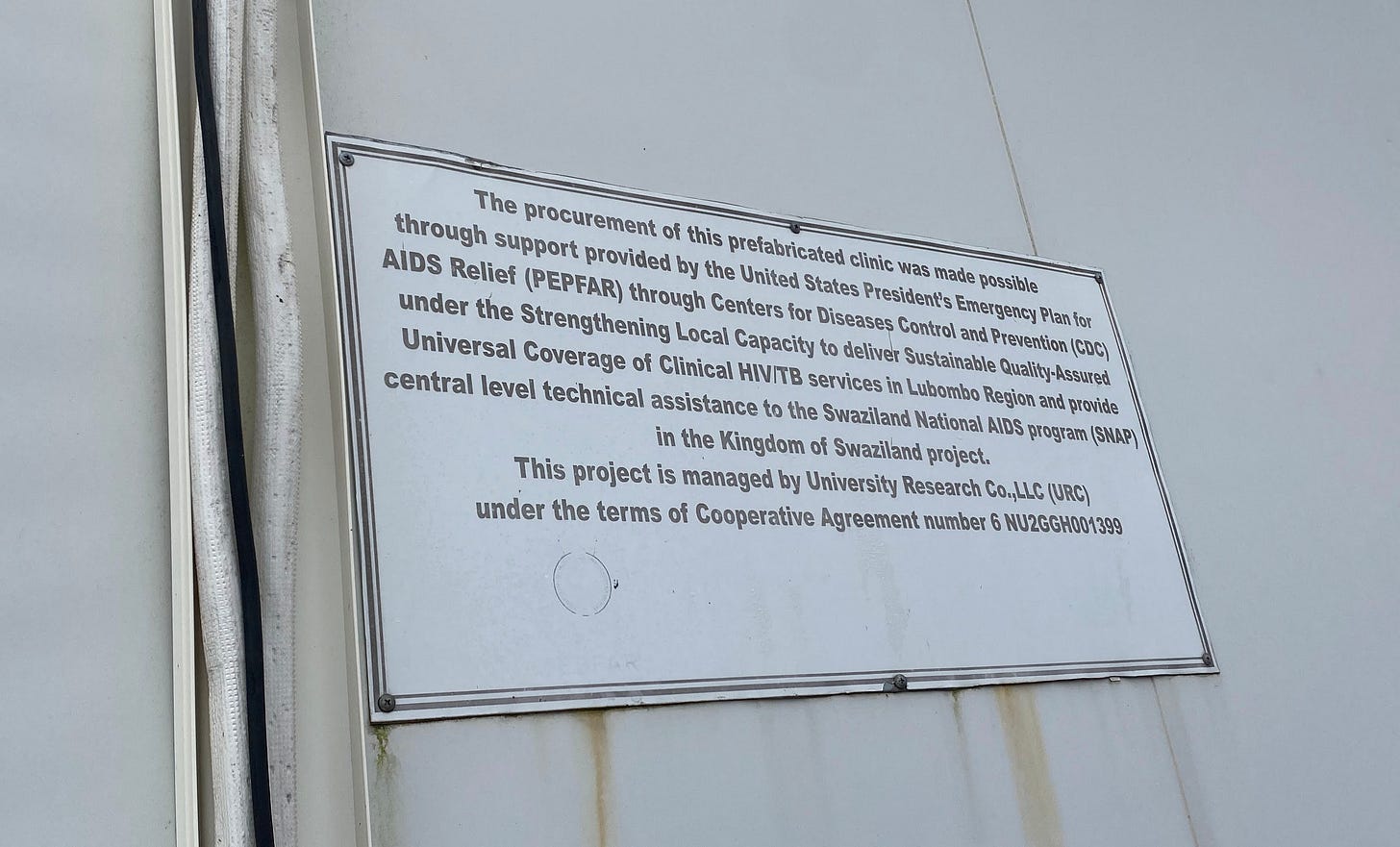 A sign on the side of a clinic in Eswatini reads, “The procurement of this prefabricated clinic was made possible through support provided by PEPFAR through CDC under the Strengthening Local Capacity to deliver Sustainable Quality-Assured Universal Coverage of Clinical HIV/TB services in Lubombo Region and provide central level technical assistance to the Swaziland National AIDS program (SNAP) in the Kingdom of Swaziland project.” At the end, it states the grant number. 