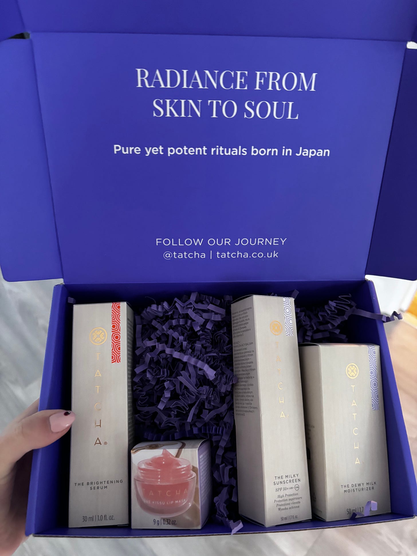 An unretouched top-down view of the royal blue Tatcha "Radiance from Skin to Soul" gift box. The unboxing features the brand's 2026 summer launches nestled in purple shredded paper, including Tatcha The Brightening Serum, Tatcha The Kissu Lip Mask, Tatcha The Milky Sunscreen SPF 50+, and Tatcha The Dewy Milk Moisturizer.