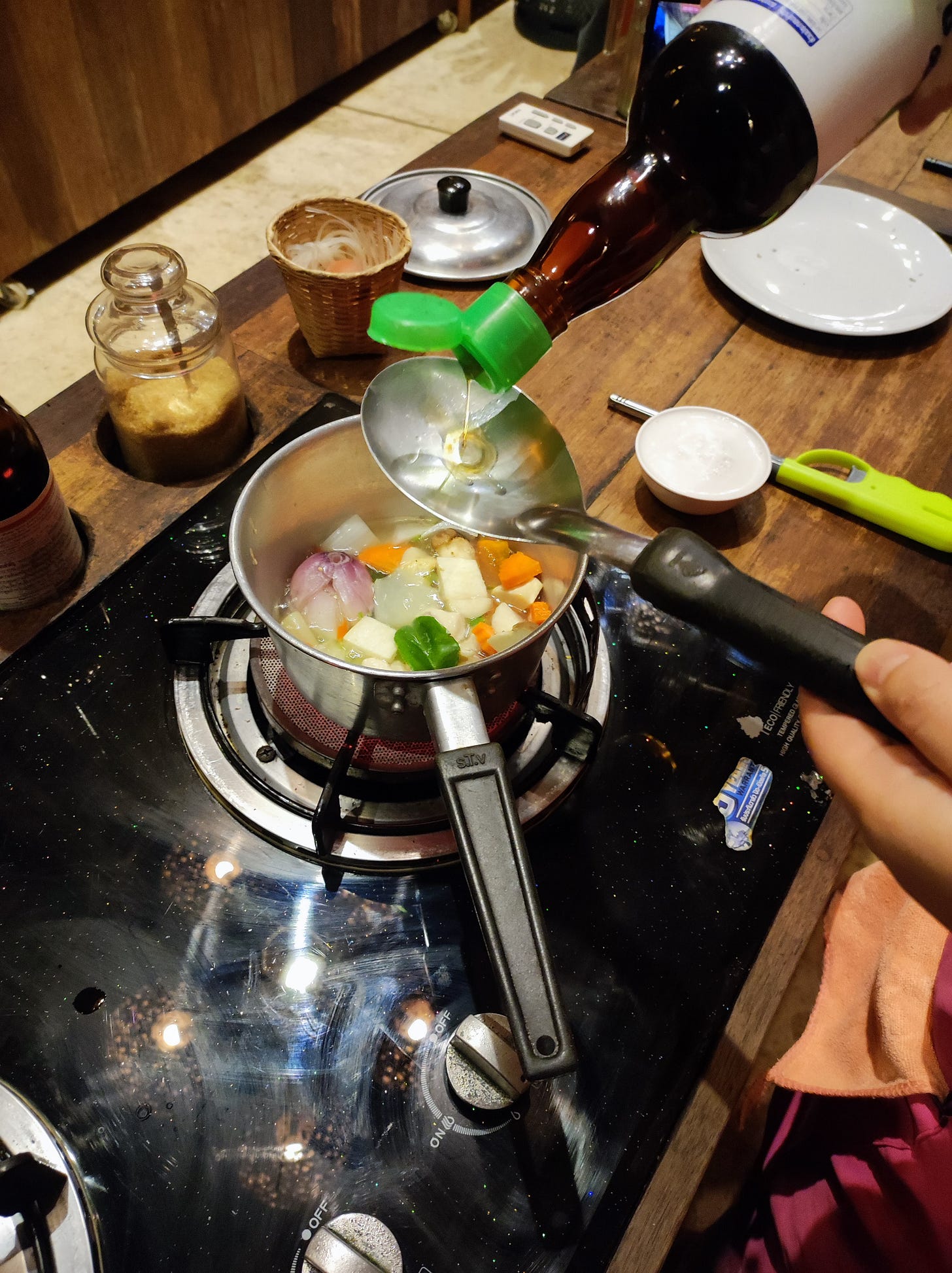 N versa una salsa in un cucchiaio, sopra una pentola che sfrigola sul fuoco. Sul bancone pieno di ingredienti.