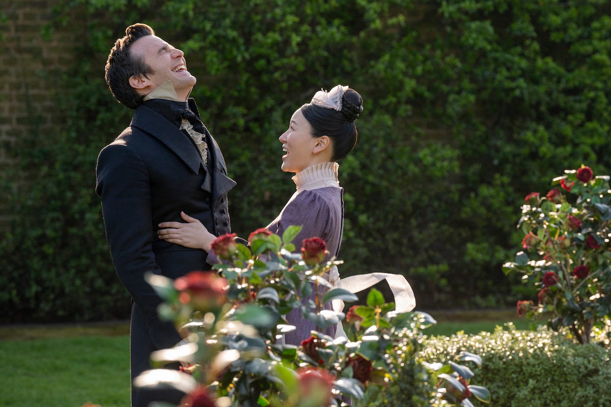 A man and woman in period costumes laugh together in a formal garden with blooming red roses and manicured greenery, under daylight. A man and woman in period costumes laugh together in a formal garden with blooming red roses and manicured greenery, under daylight.