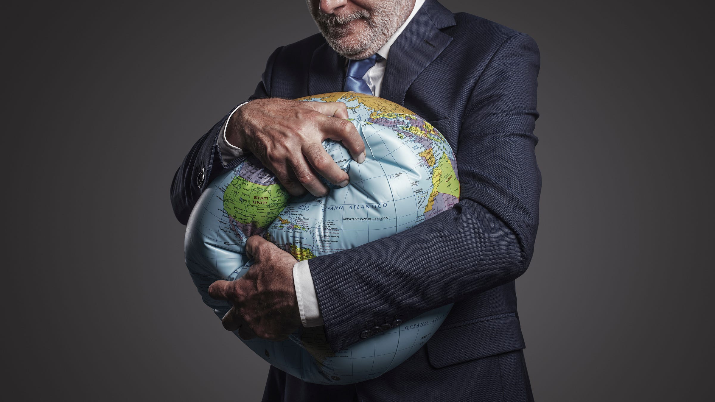 A man with a gray beard in a gray-blue suit desperately clutching an inflatable globe with both hands. 