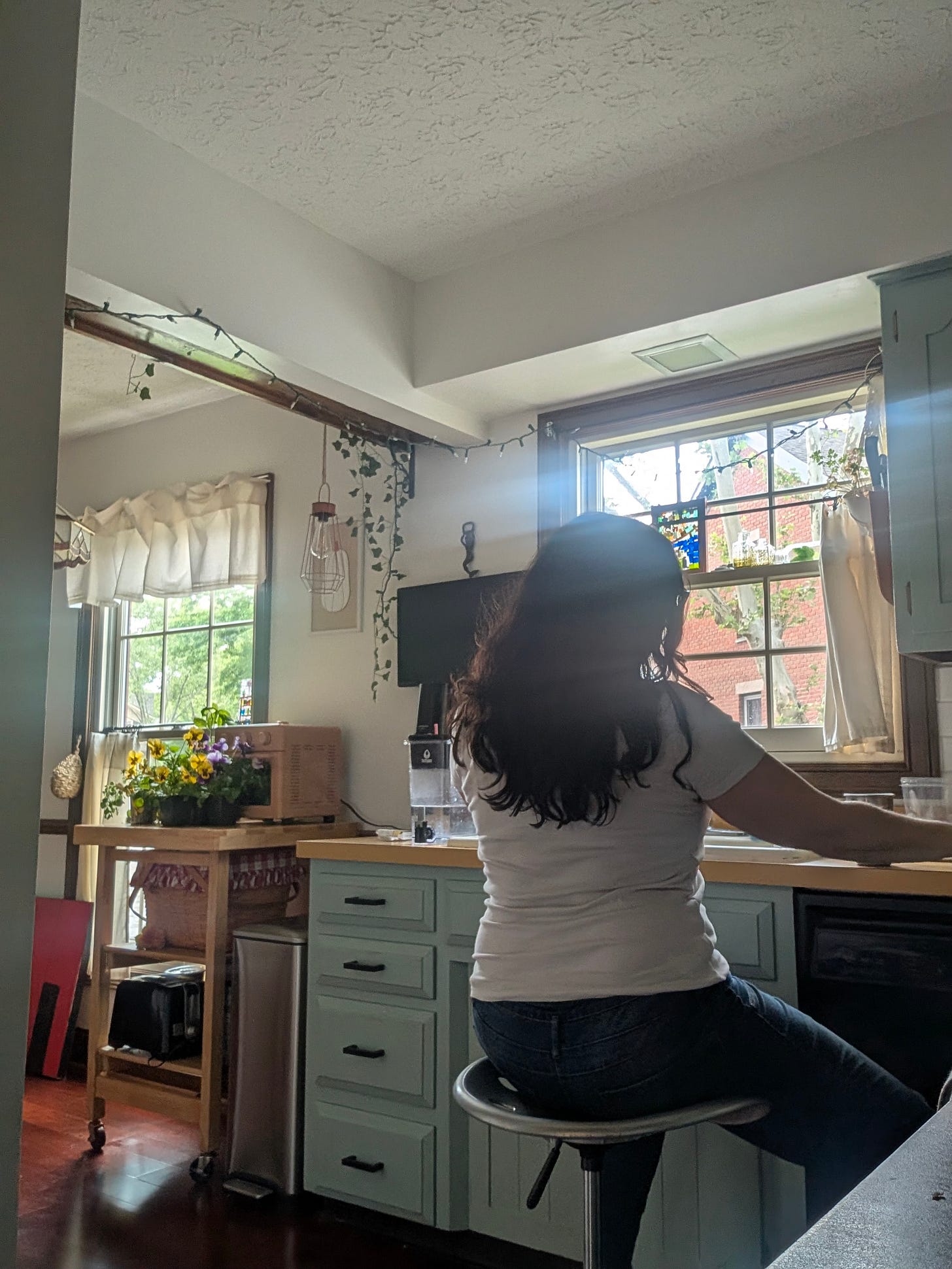 A person with long wavy dark hair, wearing a white T-shirt and jeans, sits on a silver saddle stool in a blue painted kitchen with ivy and stained glass at the windows. A person with long wavy dark hair, wearing a white T-shirt and jeans, sits on a silver saddle stool in a blue painted kitchen with ivy and stained glass at the windows.