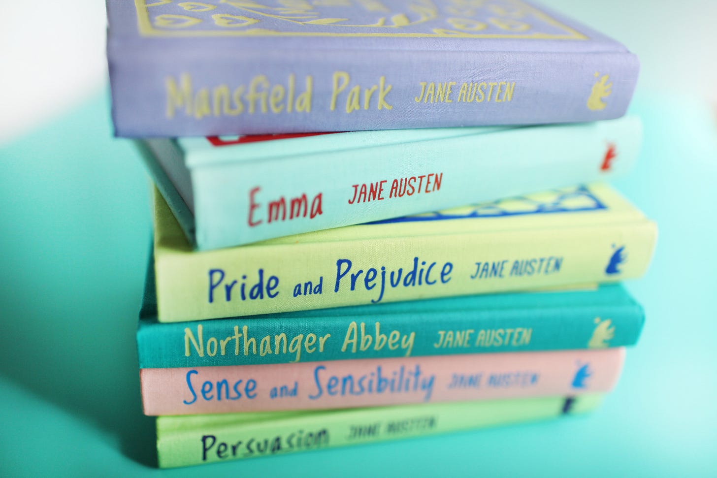 A stack of six hardcover Jane Austen novels with pastel-colored spines resting against a bright turquoise background. From top to bottom, the visible titles are Mansfield Park, Emma, Pride and Prejudice, Northanger Abbey, Sense and Sensibility, and Persuasion.