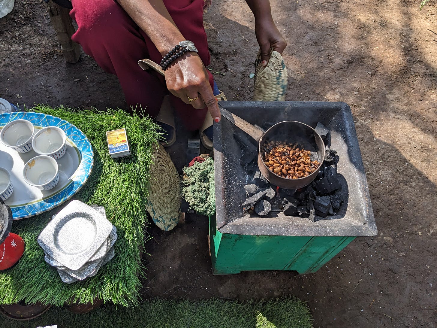 A black woman, wearing a red outfit, is kneeling down, roasting coffee over an open fire. We don’t see her face. A black woman, wearing a red outfit, is kneeling down, roasting coffee over an open fire. We don’t see her face.