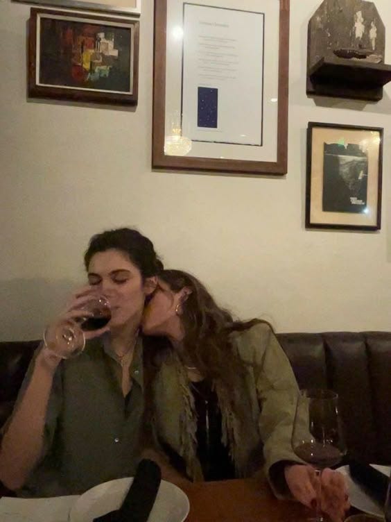 This may contain: two women sitting at a table with wine glasses in front of their faces, one woman is kissing the other