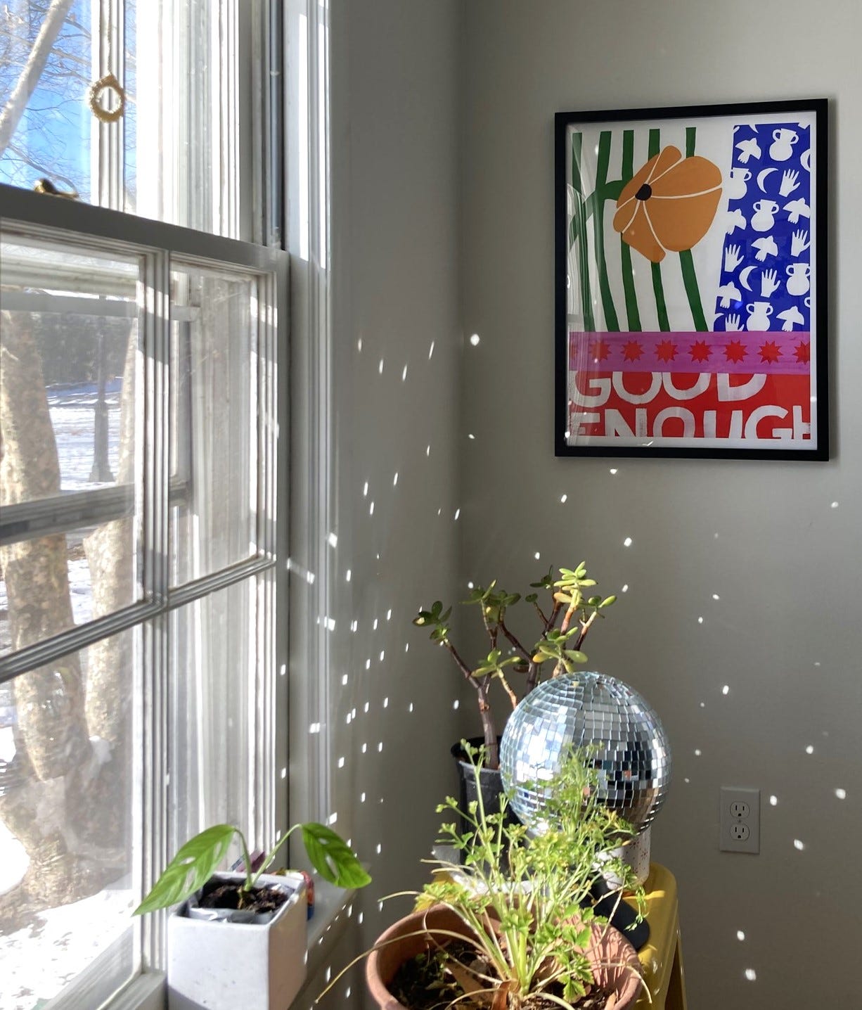 Sun streaming through my dining room window. There's a blue sky outside and snow on the ground. AN Anisa Makhoul poster is framed on the wall and reads GOOD ENOUGH in words that are legible but intentionally cut off. Some plants sit on a stool in the foreground, along with a disco ball that's casting prismatic light around the corner of the room. Sun streaming through my dining room window. There's a blue sky outside and snow on the ground. AN Anisa Makhoul poster is framed on the wall and reads GOOD ENOUGH in words that are legible but intentionally cut off. Some plants sit on a stool in the foreground, along with a disco ball that's casting prismatic light around the corner of the room.