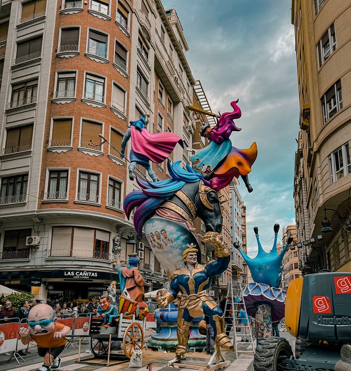 A gallery of Fallas monuments in Valencia, Spain — elaborately sculpted figures including a Charlie Chaplin figure holding butterflies, fantasy warriors, and colorful street sculptures, photographed against dramatic cloudy and blue skies in March 2026.