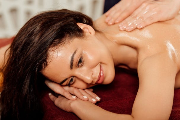 A woman with dark hair is receiving a shoulder massage. She is smiling gently and appears relaxed. Her skin has a slight sheen, possibly from massage oil. The background is blurred, focusing attention on her face and the hands massaging her shoulder.