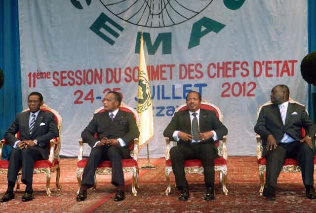 Four men in dark suits, seated in front of a large banner