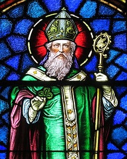 Stained glass image of St. Patrick. The saint is depicted wearing mostly green with white robes, holding a shamrock, the background glass is blue. 