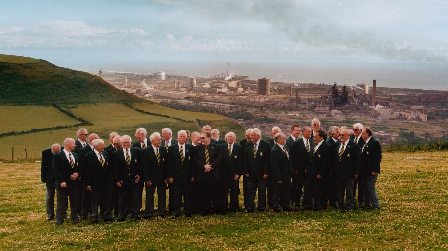 A male voice choir in dark green suits, white shirts and green and yellow striped ties standing on a green hill with yellow daisies. In the background is the industrial town of Port Talbot and the large steelworks. To the left of the frame is another lush green hillside. In the distance is the sea and a blue sky with little cloud and some grey smoke. A male voice choir in dark green suits, white shirts and green and yellow striped ties standing on a green hill with yellow daisies. In the background is the industrial town of Port Talbot and the large steelworks. To the left of the frame is another lush green hillside. In the distance is the sea and a blue sky with little cloud and some grey smoke.