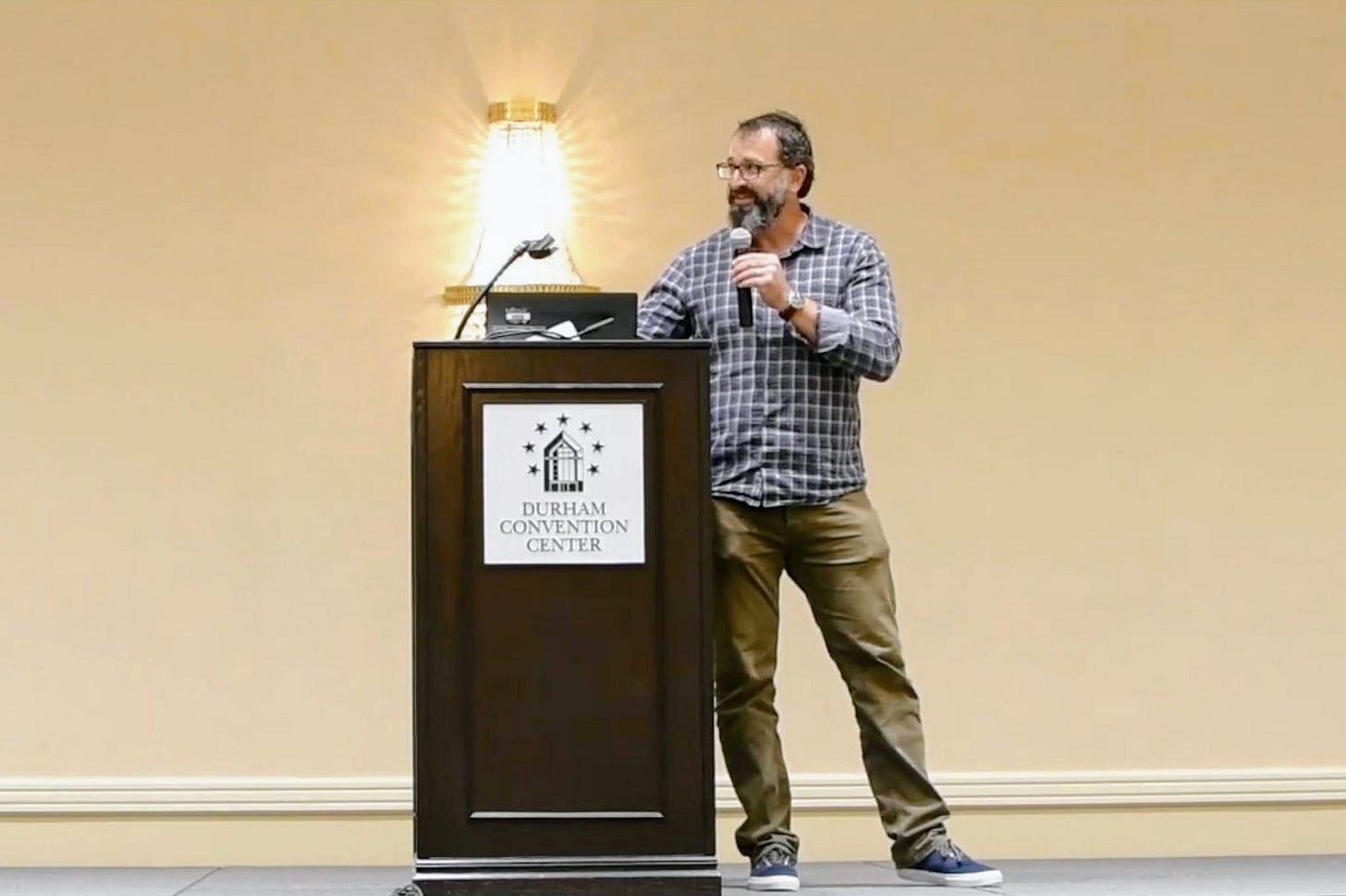 The author standing at a wooden podium at the Durham Convention Center, holding a microphone and speaking. He wears glasses, a gray beard, a blue plaid button-down shirt, khaki pants, and blue sneakers. A wall sconce glows behind the podium. The author standing at a wooden podium at the Durham Convention Center, holding a microphone and speaking. He wears glasses, a gray beard, a blue plaid button-down shirt, khaki pants, and blue sneakers. A wall sconce glows behind the podium.
