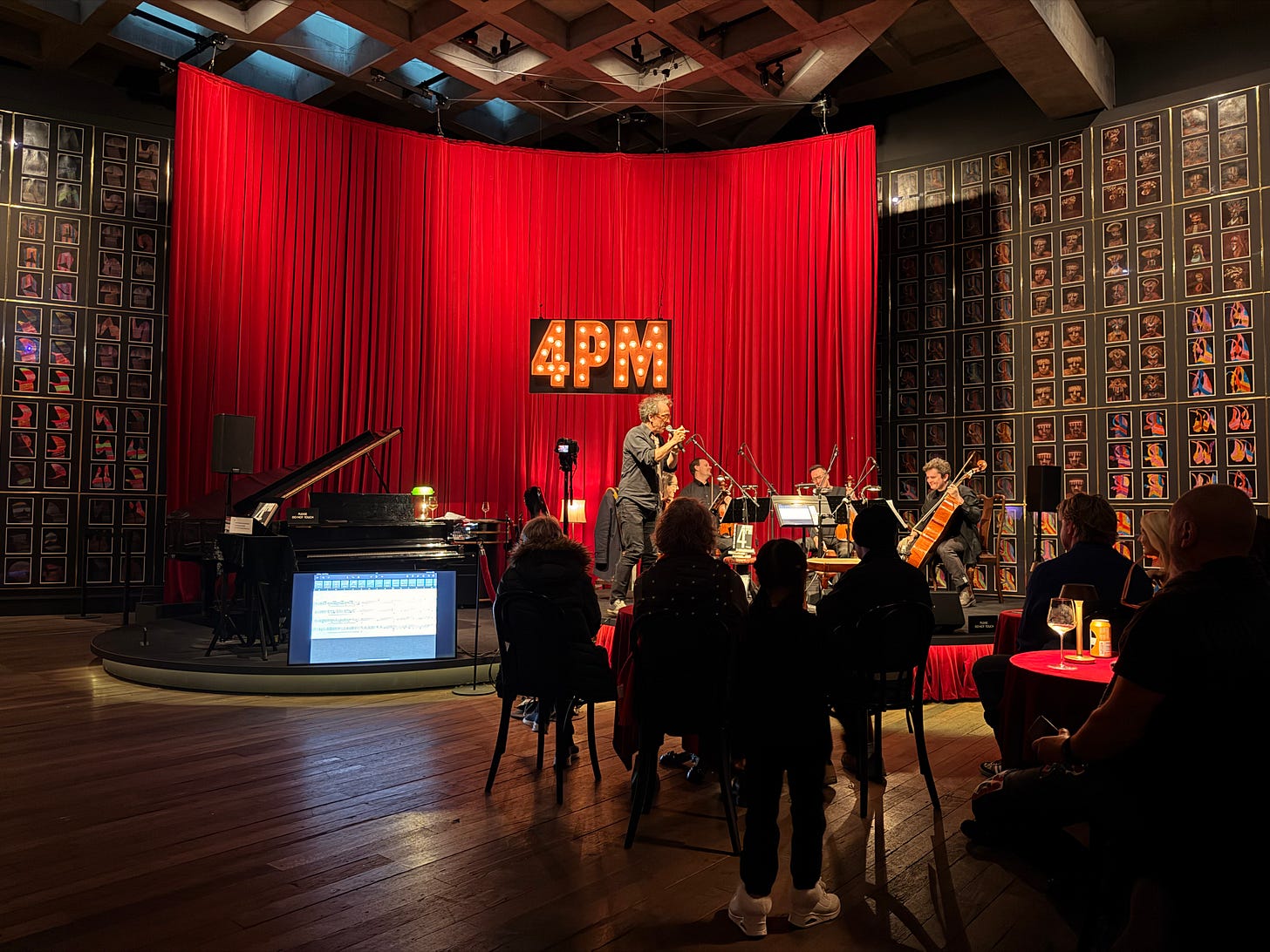 Musicians performing live at MONA with a red curtain backdrop and audience seated closely around them.