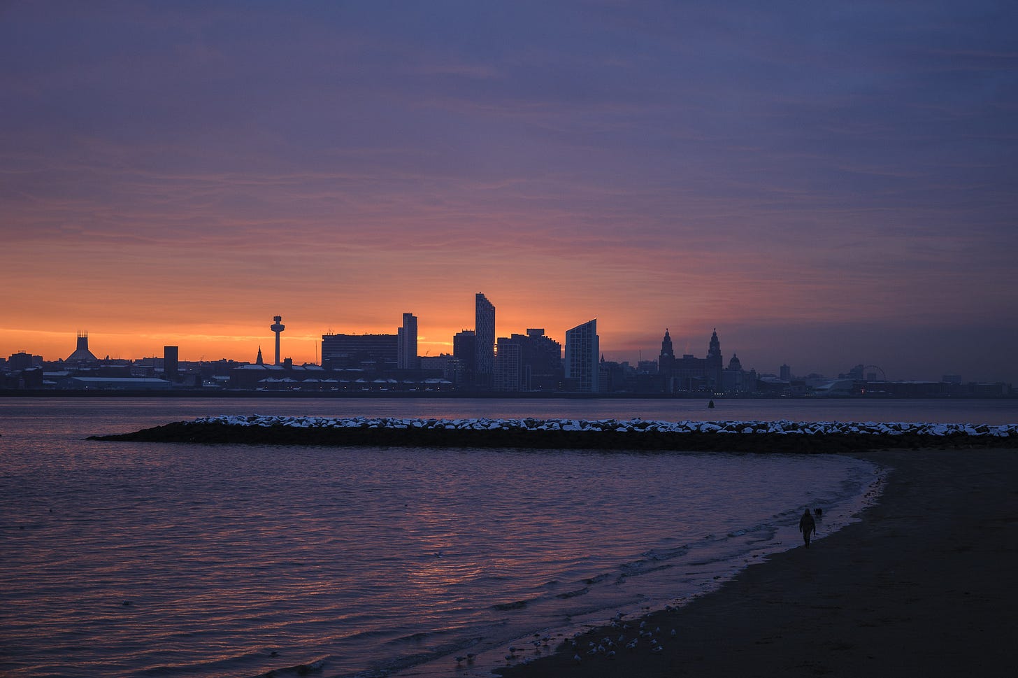 As the sky turns purple a person walks along the river bed of the Mersey while the tide is out. It’s a cold morning and there is a dusting of snow on the rocks.