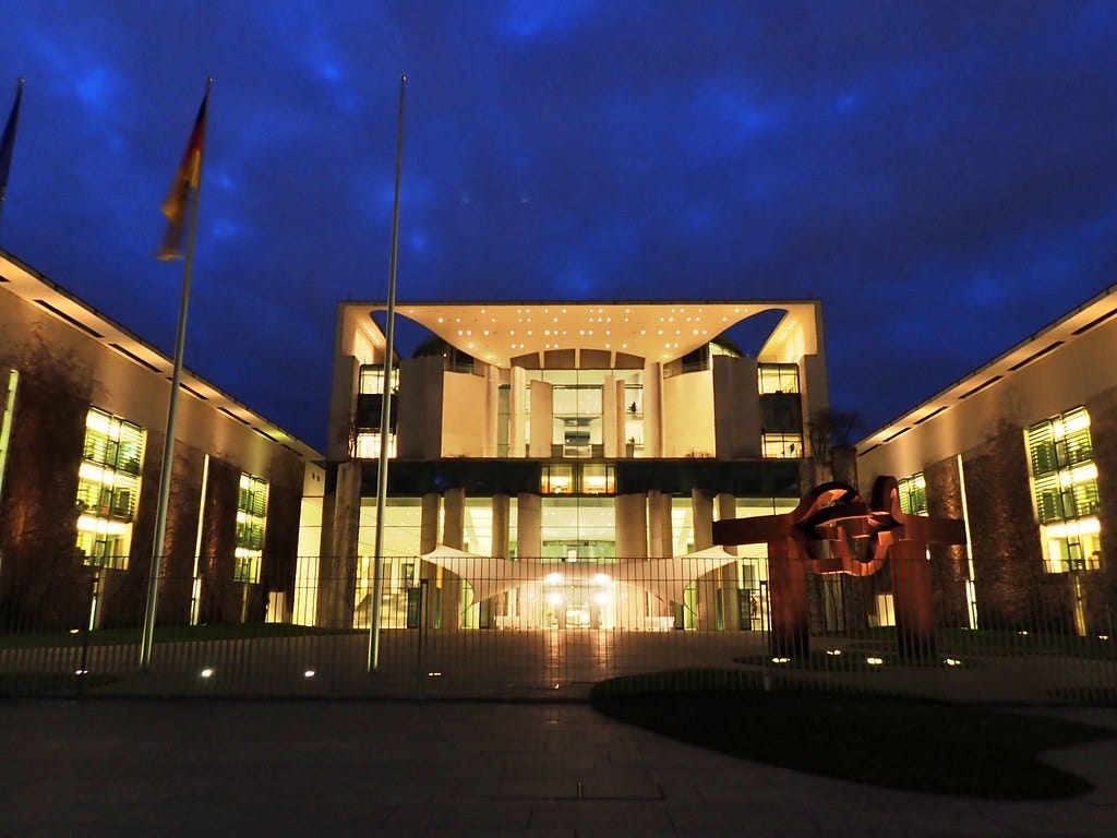 Illuminated modern government building at night, with German flags and a gated forecourt. Illuminated modern government building at night, with German flags and a gated forecourt.