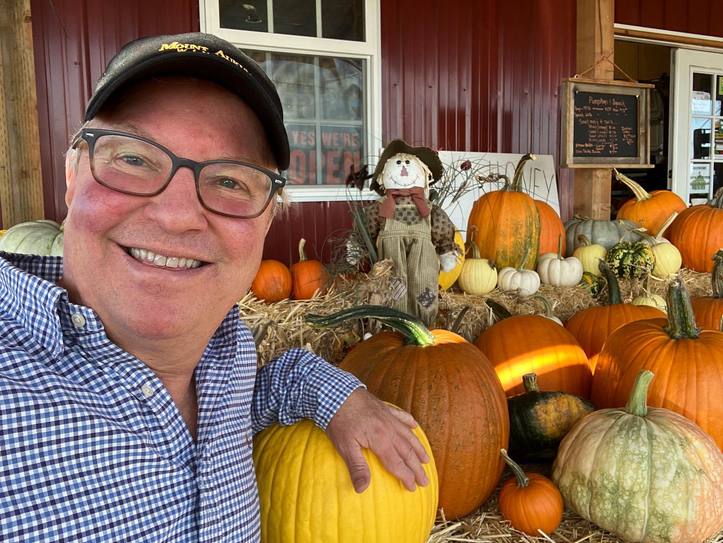 Man wearing hat and glasses, posing by pumpkins and a scarecrow