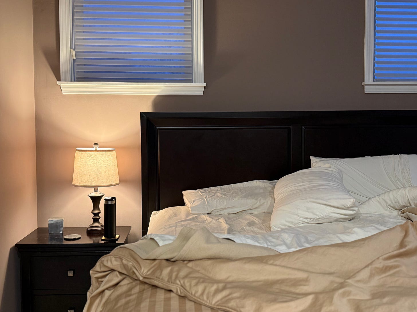 A rumpled bed in soft early-morning light, bedside lamp glowing warm against cool window light, with a dark water bottle on the nightstand — the quiet moment before the day begins.