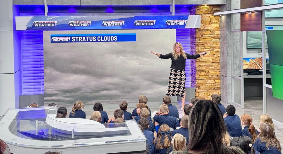 A woman in a black blouse and checkered skirt stands gesturing in front of a large weather screen displaying stratus clouds inside a modern TV studio with blue lighting and weather channel branding. Behind her a group of young children in school uniforms sit attentively on the floor listening. The setting includes studio equipment like a green screen and wooden accents.