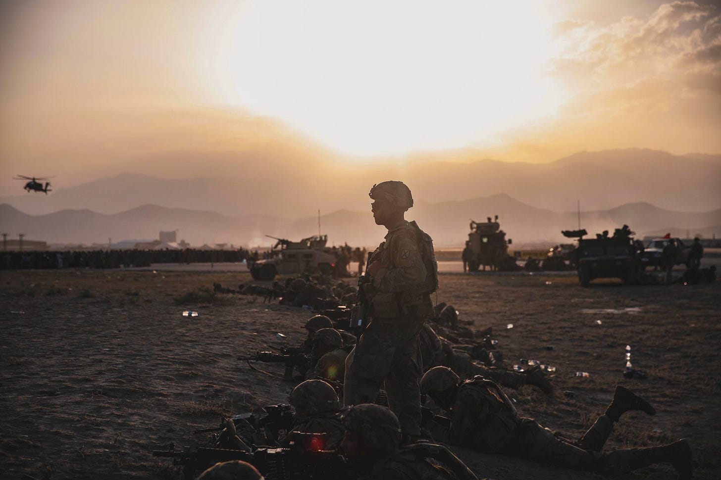 A soldier stands in the middle of the image facing left. Other soldiers are lying flat on the ground in a line alongside him. The sky is bright and dusty, and there are mountains visible on the horizon. Various military equipment, including what appear to be tanks, humvees, and a helicopter, are visible in the background. A soldier stands in the middle of the image facing left. Other soldiers are lying flat on the ground in a line alongside him. The sky is bright and dusty, and there are mountains visible on the horizon. Various military equipment, including what appear to be tanks, humvees, and a helicopter, are visible in the background.