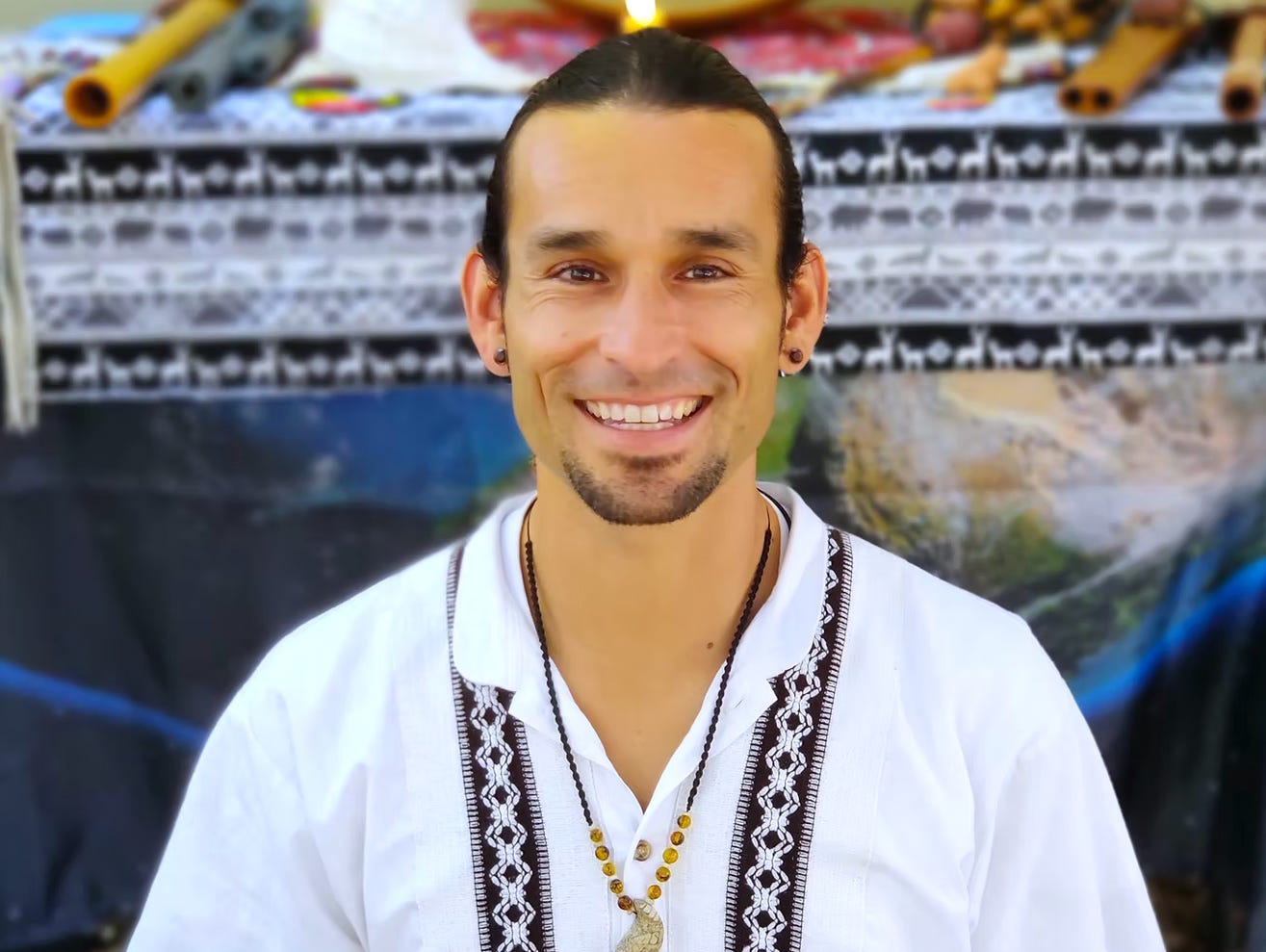 Gera, a young man with his hair pulled back and a small beard, smiles brightly at the camera. He wears a button down white shirt with black embroidery. Behind him is an altar with a blue and white and black cloth and various musical instruments.