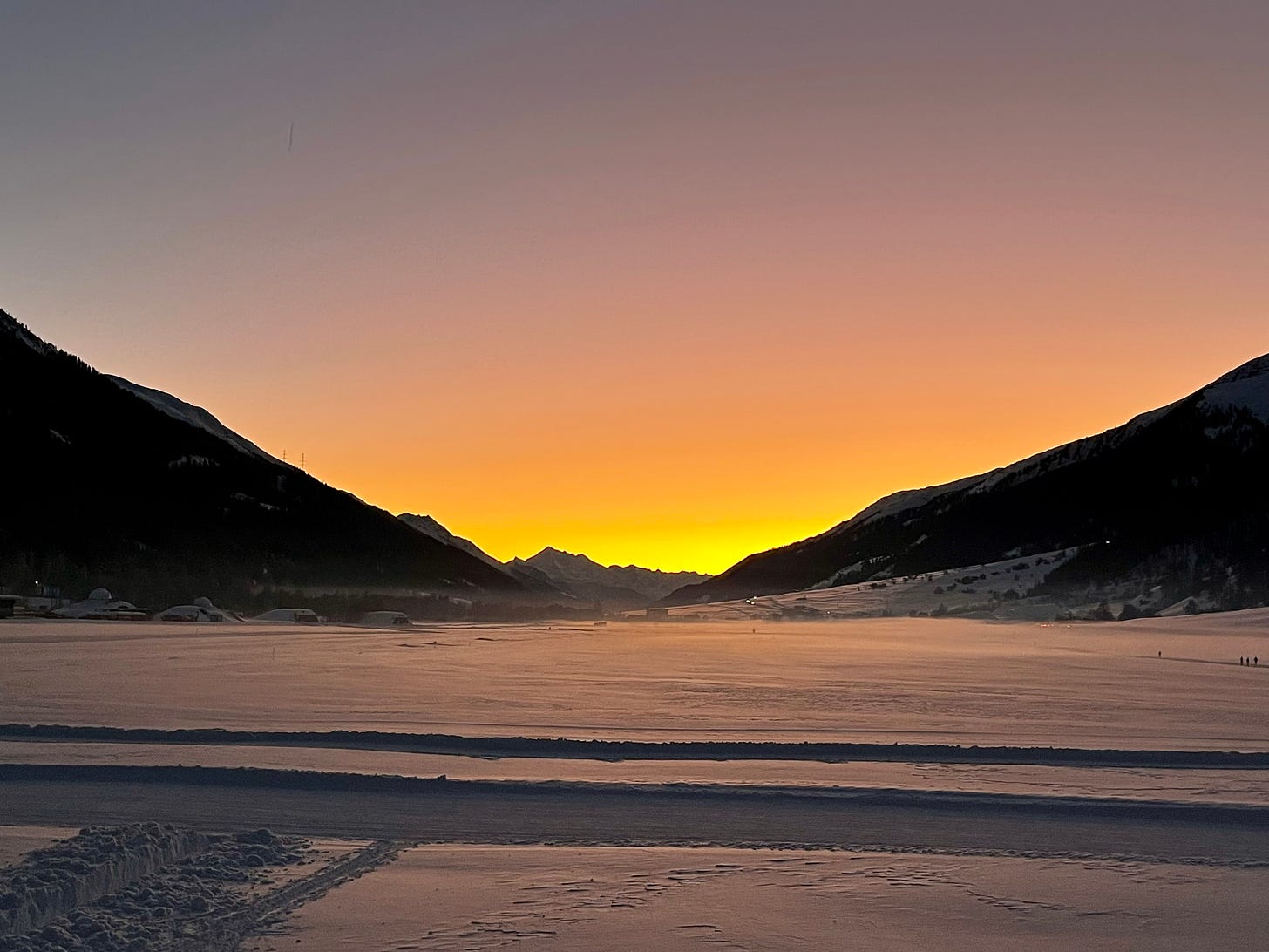 A snow covered valley bordered by tree-covered mountain slopes that frame the Weisshorn, one of Switzerland's highest peaks. The snow glows golden and sky goes from a bright yellow to orange to pink to blue.