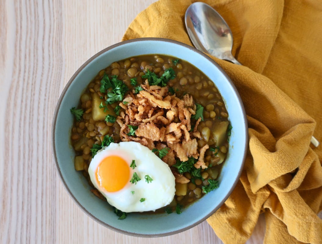 A bowl of adasi lentil soup topped with parsley and egg, with a yellow napkin and a gold spoon.