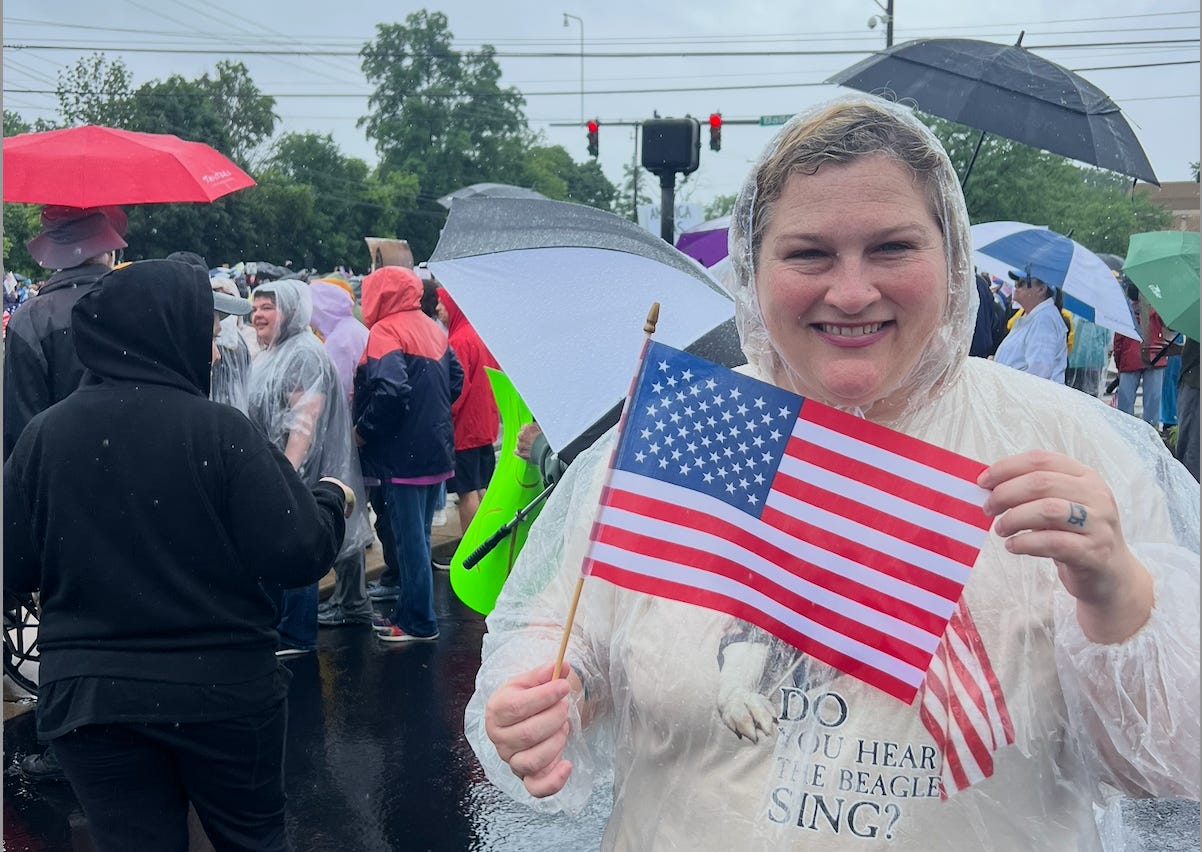 Jamie smiles in a rain poncho holding a small American flag