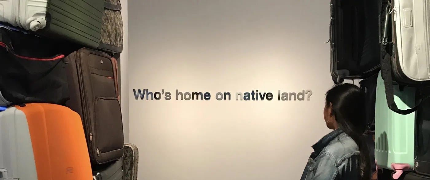 Justine's back is to the camera as she faces a sign that reads "Who's home on native land". All around her are suitcases. 