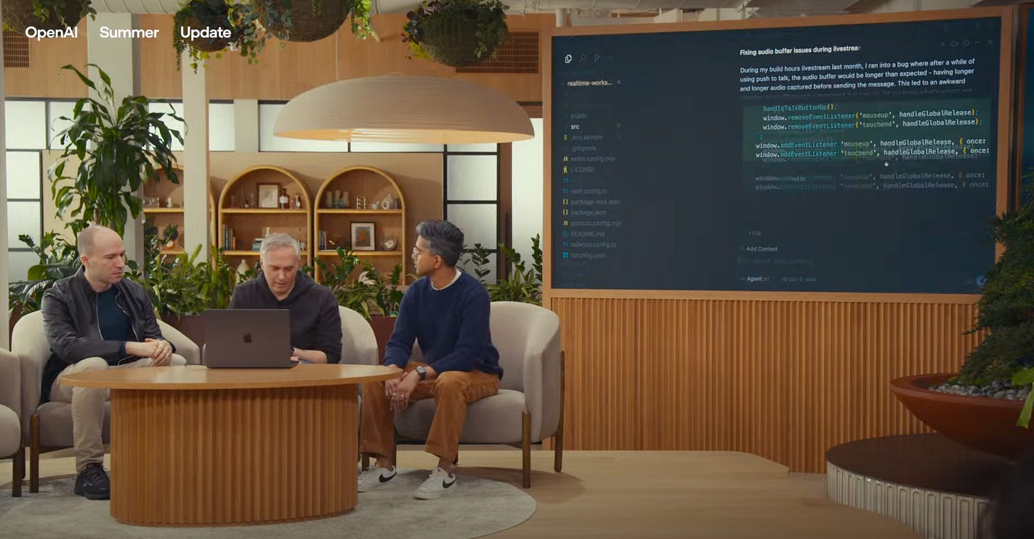 Still from the OpenAI GPT-5 launch event, showing three people sat around a laptop as code is displayed on a large screen behind them