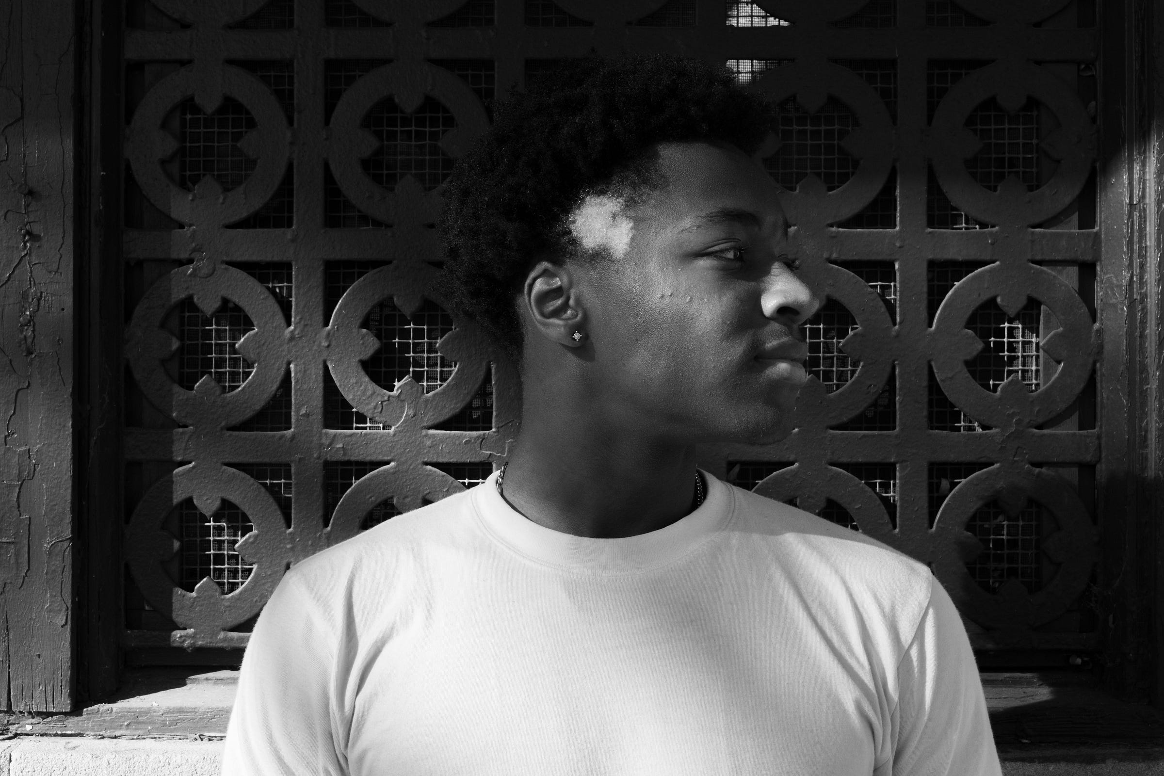 A black-and-white portrait of a young man wearing a white t-shirt, standing in front of an ornate, patterned metal grate. He gazes to the side with a calm yet contemplative expression, the sunlight casting soft shadows on his face. A distinct patch of lighter hair near his temple adds contrast and character. The background features a textured wall with peeling paint, emphasizing the contrast between structure and individuality.