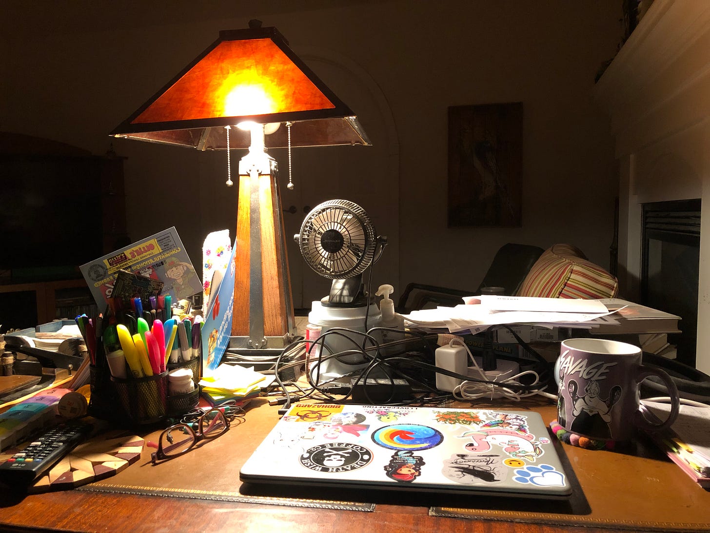 The author's desk featuring a laptop, a pen caddy, glasses, coffee mug, and mission-style lamp