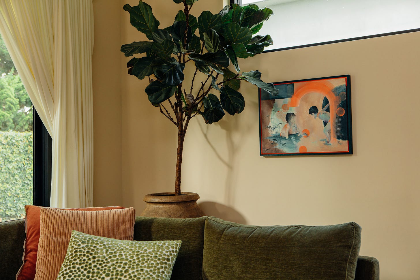 Living room corner with a fiddle-leaf fig in a terracotta pot, a green velvet sofa with coral and peach cushions, and Assia Bennani's Playtime, a painting of two children in water under an orange arch, hung on a warm beige wall.