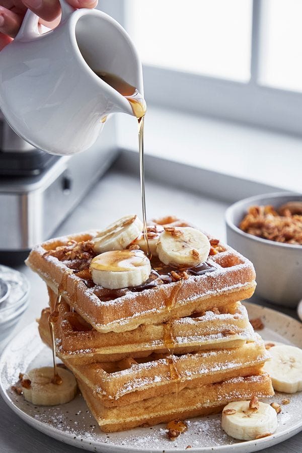 This may contain: a stack of waffles with bananas and syrup being poured over them on a plate