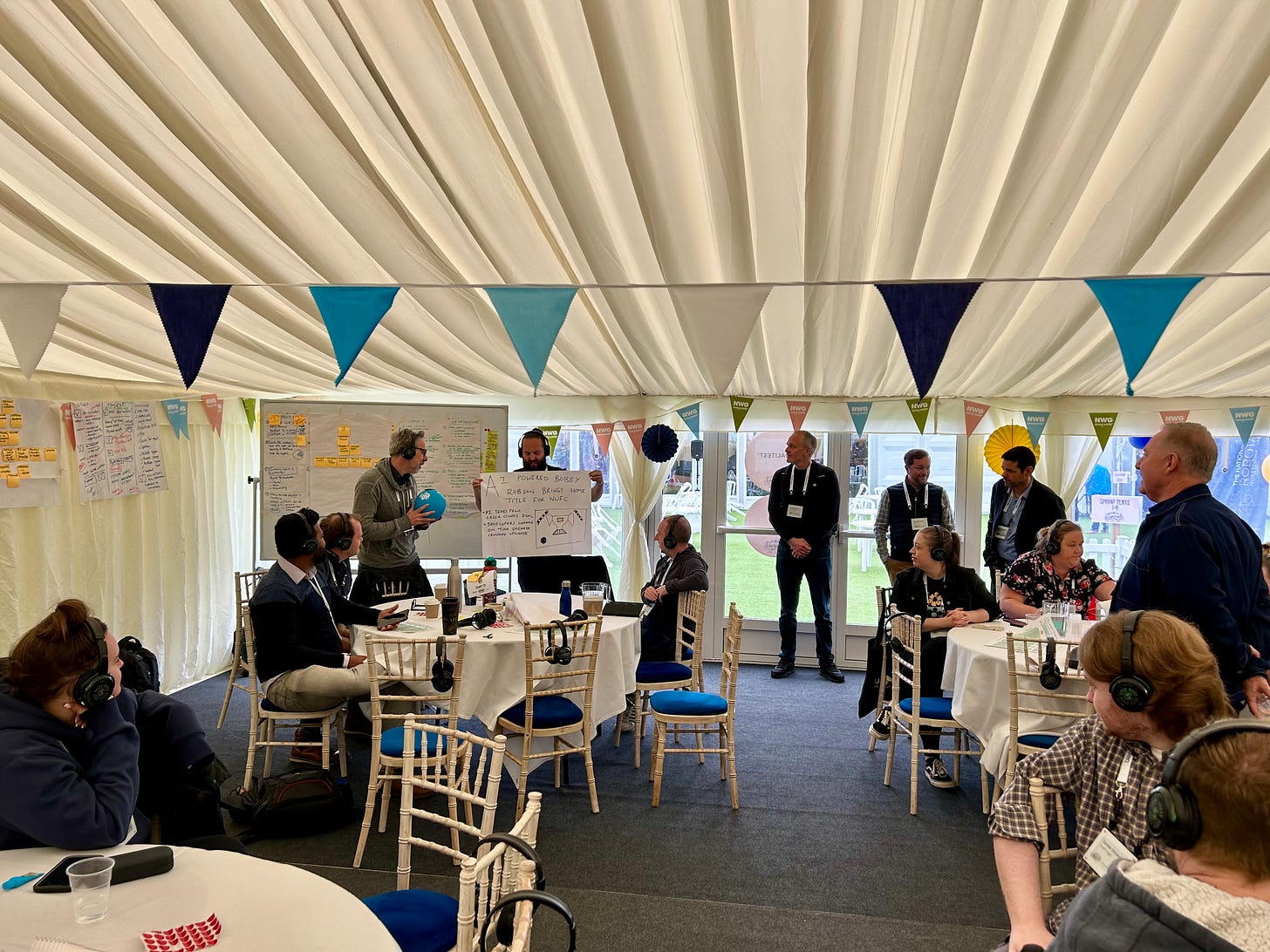 A workshop in full swing. A man holds a blue ball that is a microphone and describes a newspaper front page from the future that his team mate holds up for everyone to see. People stand and sit around watching. There are white boards covered in post it notes in the background.