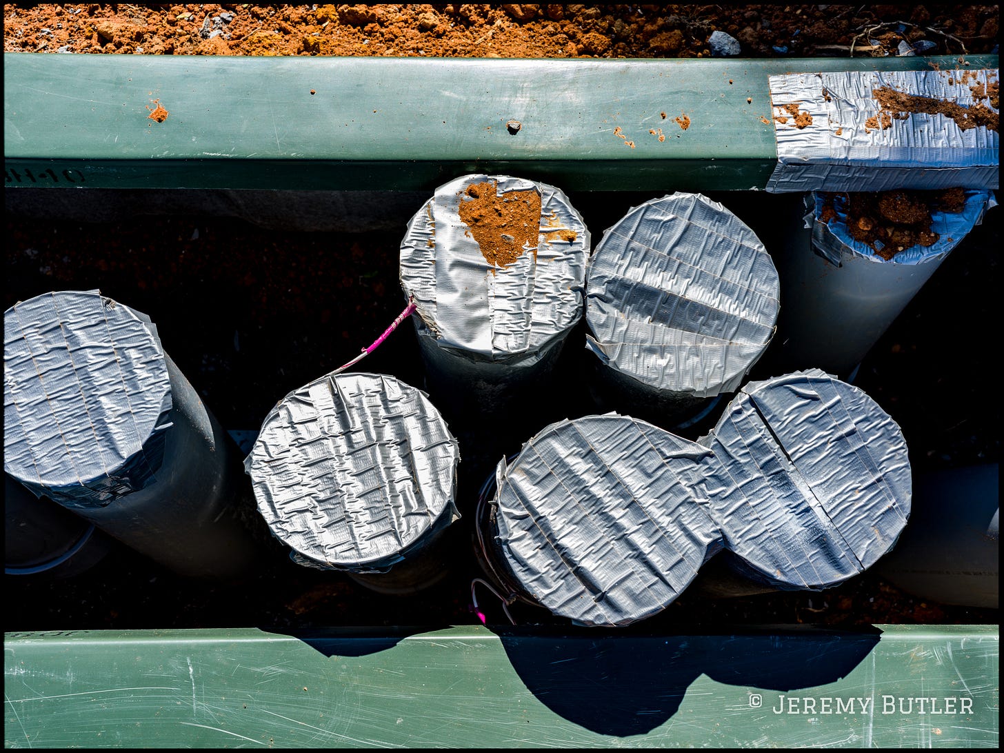 A top-down view of several pipes covered with duct tape at a construction site. The pipes are placed alongside a green barrier and surrounded by exposed earth. A top-down view of several pipes covered with duct tape at a construction site. The pipes are placed alongside a green barrier and surrounded by exposed earth.