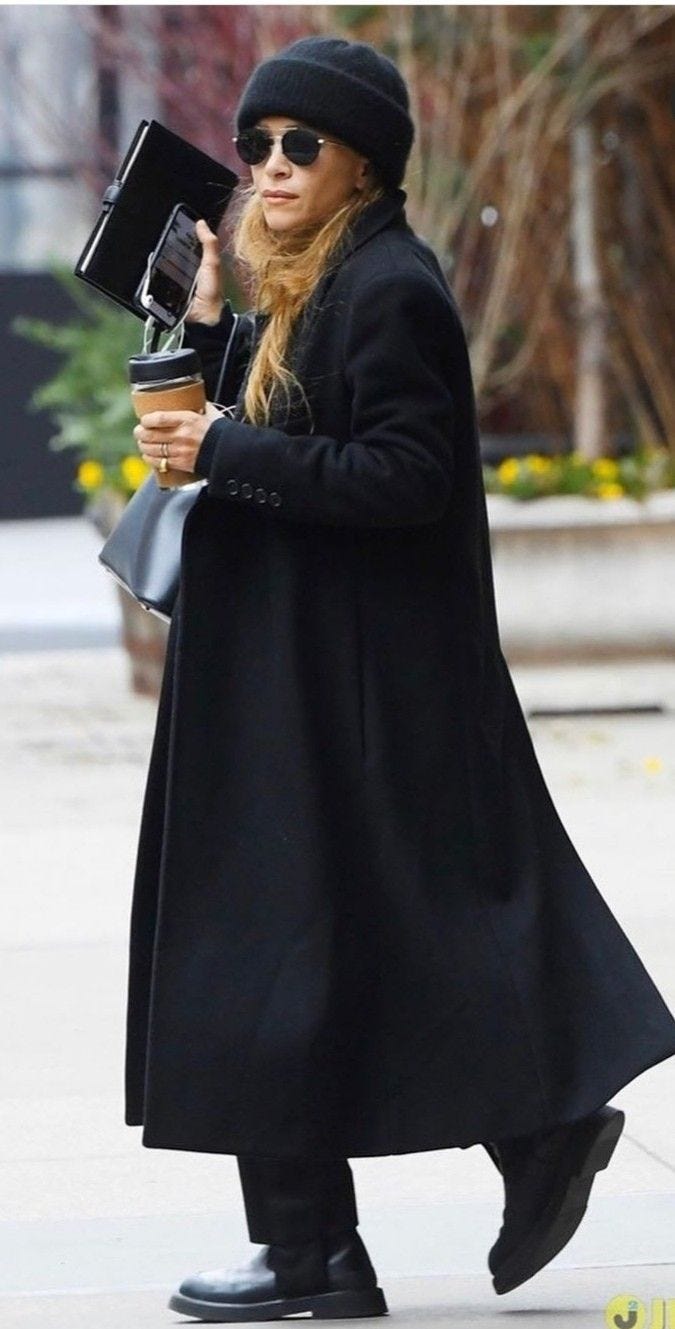 This may contain: a woman in black coat and hat walking down the street with a camera on her hand