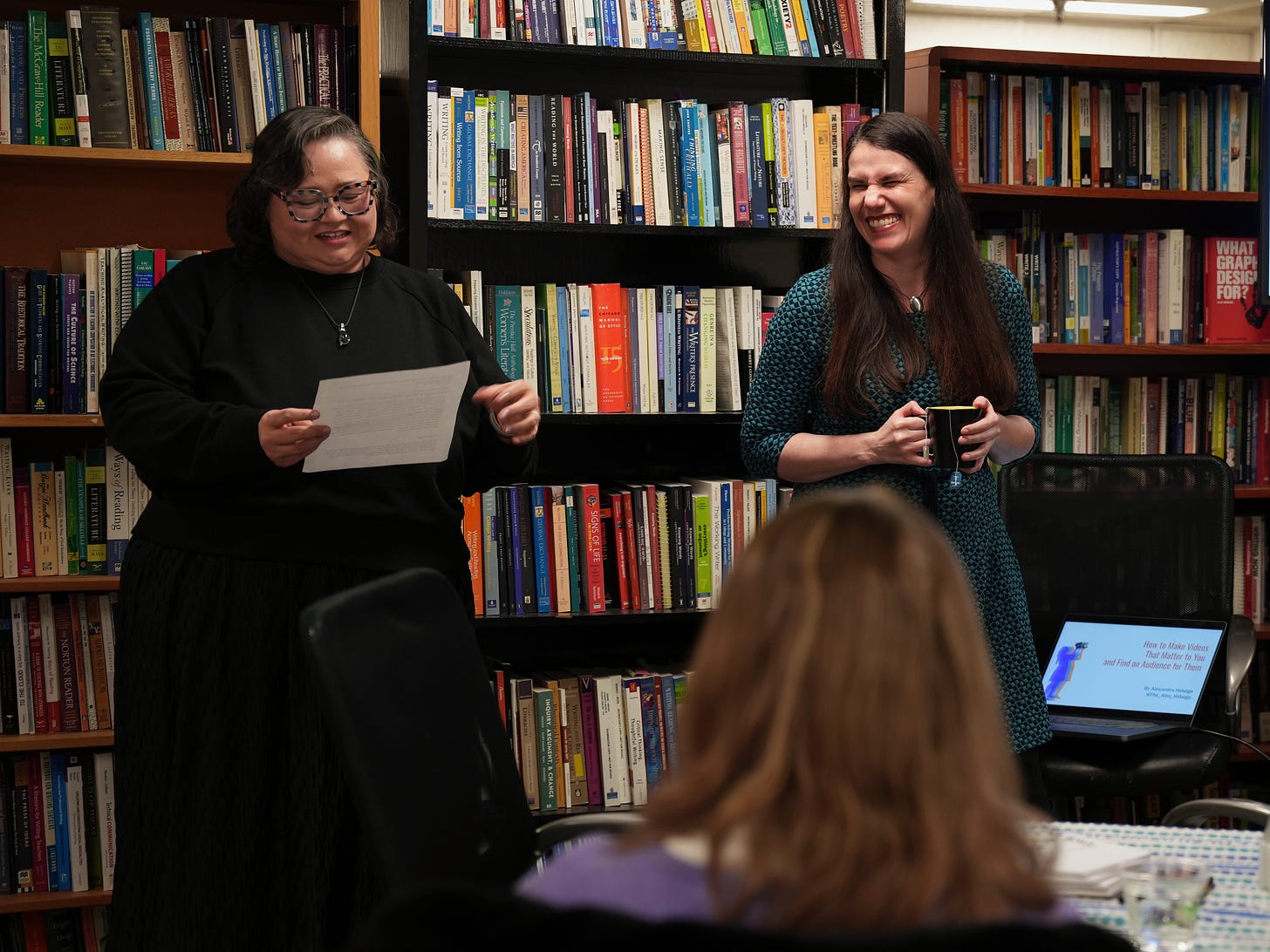 Two women stand in a room with many bookshelves behind them. One (Jaquetta Shade) is holding a paper and reading from it and smiling as she does. The other (Alexandra Hidalgo) is laughing as she holds a mug of tea. We see a computer and a TV presentation screen to the side. We also see the back of a woman's head in the front. The woman is in the audience. It is daytime. They are in a conference room at The University of Colorado Boulder. Two women stand in a room with many bookshelves behind them. One (Jaquetta Shade) is holding a paper and reading from it and smiling as she does. The other (Alexandra Hidalgo) is laughing as she holds a mug of tea. We see a computer and a TV presentation screen to the side. We also see the back of a woman's head in the front. The woman is in the audience. It is daytime. They are in a conference room at The University of Colorado Boulder.