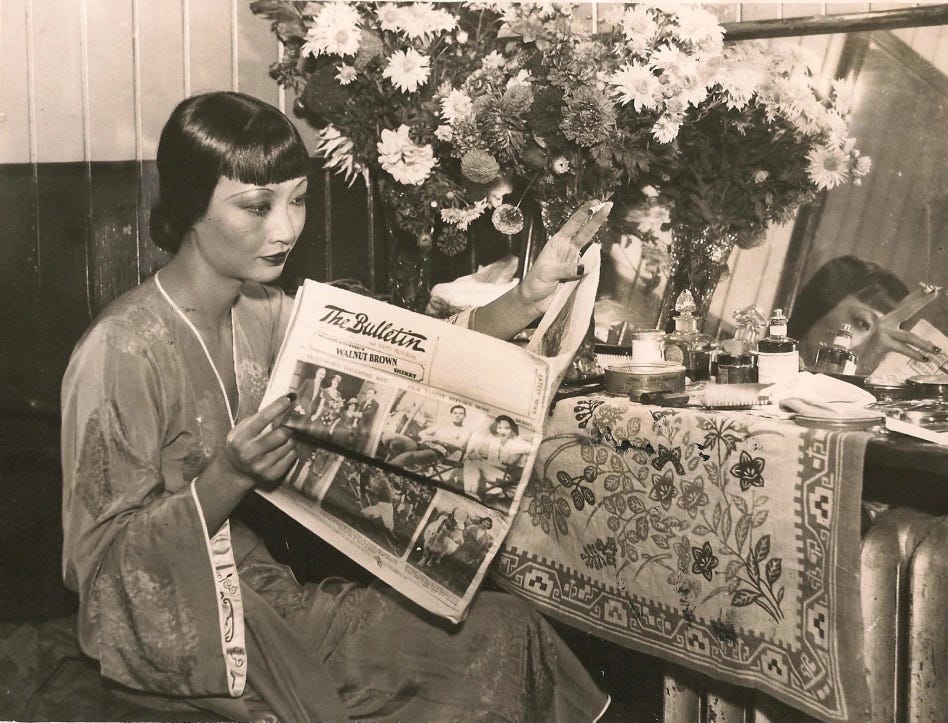 Anna May Wong in a silk robe sits at her backstage dressing table strewn with bouquets and beauty products, she reads The Bulletin newspaper held in her hands Anna May Wong in a silk robe sits at her backstage dressing table strewn with bouquets and beauty products, she reads The Bulletin newspaper held in her hands