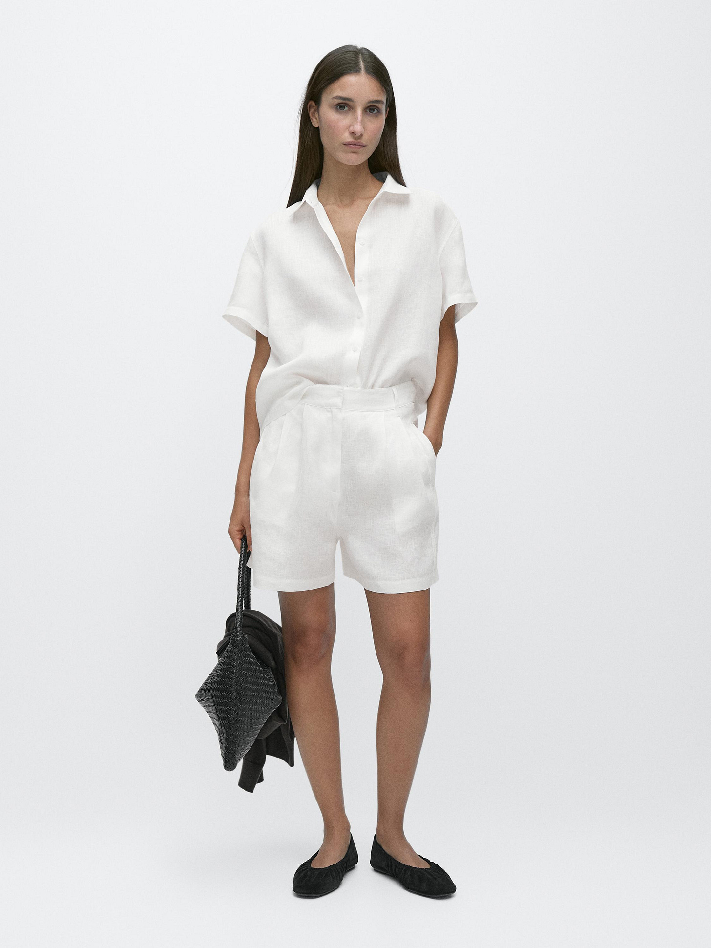Short-sleeved white shirt and long Bermuda shorts with pleats, side pockets. Wears a black bag and black flat shoes.