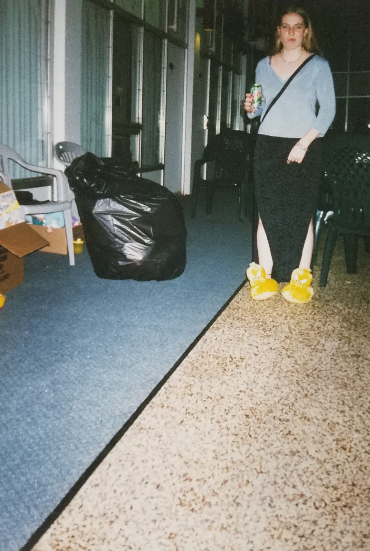 A pale teenager with long dark blong hair stands on a pebbled floor near the doors of many hotel rooms that look out onto the indoor pool area. They are wearing a light blue v-neck shirt and a black maxi skirt with slits up both sides to the knee and blue flowers that match the shirt. On their feet are Tweety bird slippers. They are holding a can of Mt Dew and looking dazed (not at the camera) as it appears they are biting the inside of their cheek in this candid moment that someone captured on film. 