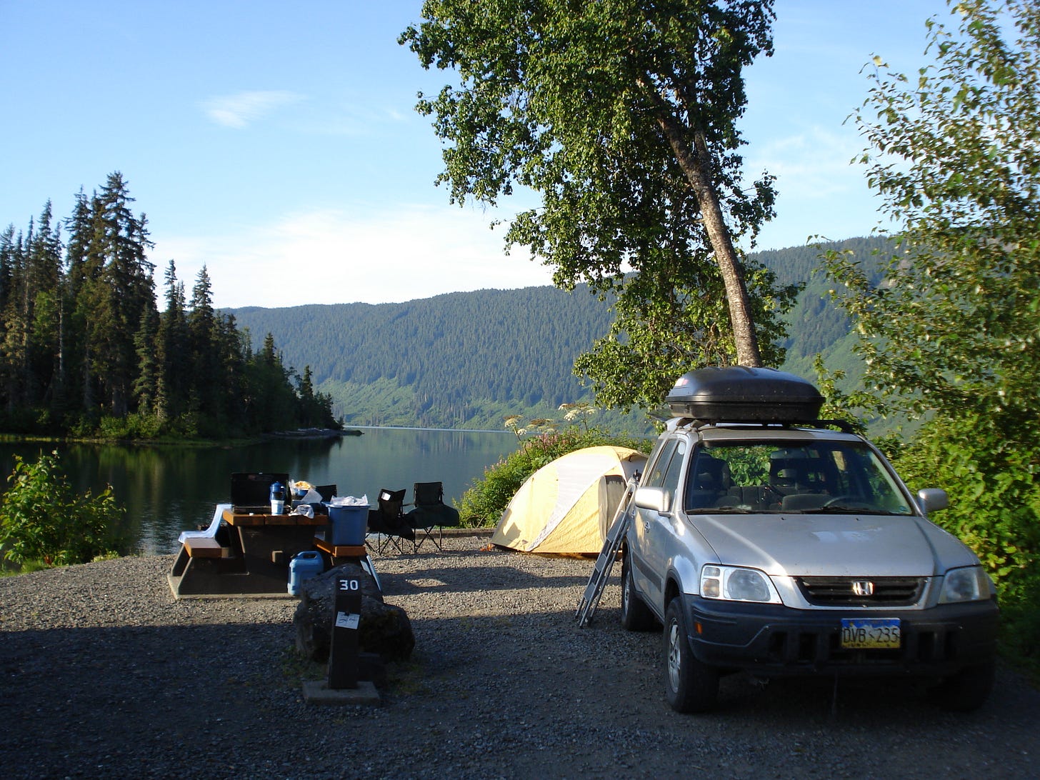 A silver Honda CRV sits in front of a wooded lake with a tent and camping gear set up behind it.