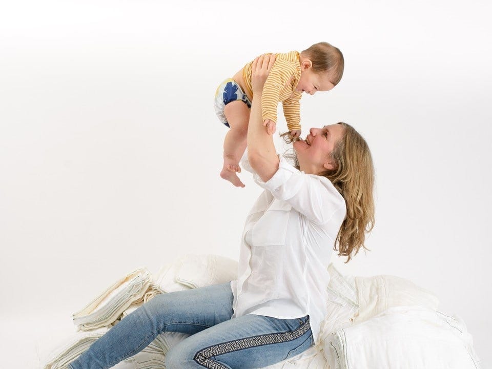 A white woman with blond shoulder-length hair wearing a white blouse smiles at the brown-haired baby she holds over her head. A white woman with blond shoulder-length hair wearing a white blouse smiles at the brown-haired baby she holds over her head.