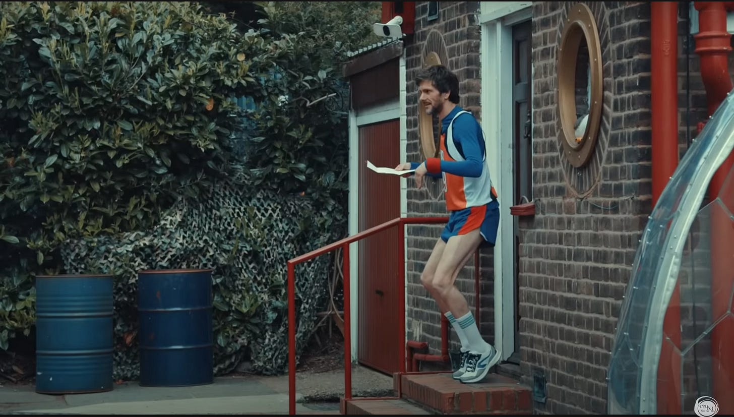 Mathew Baynton, wearing a singlet and short shorts, assumes a tiptoe position.