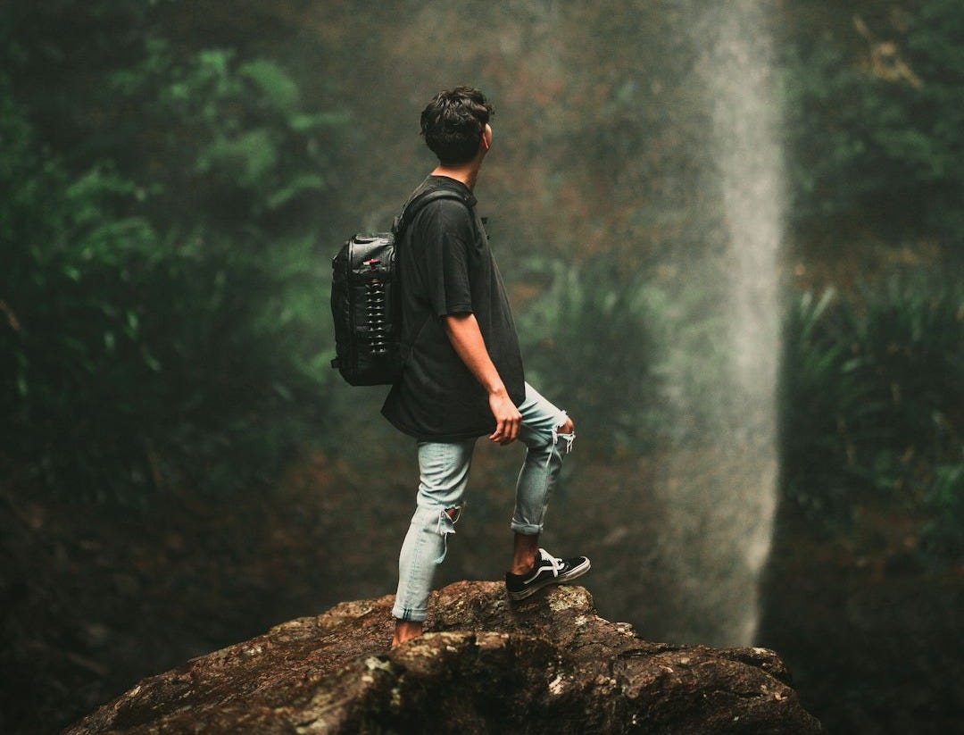 man in black t-shirt and blue denim jeans standing on rock near waterfalls during daytime