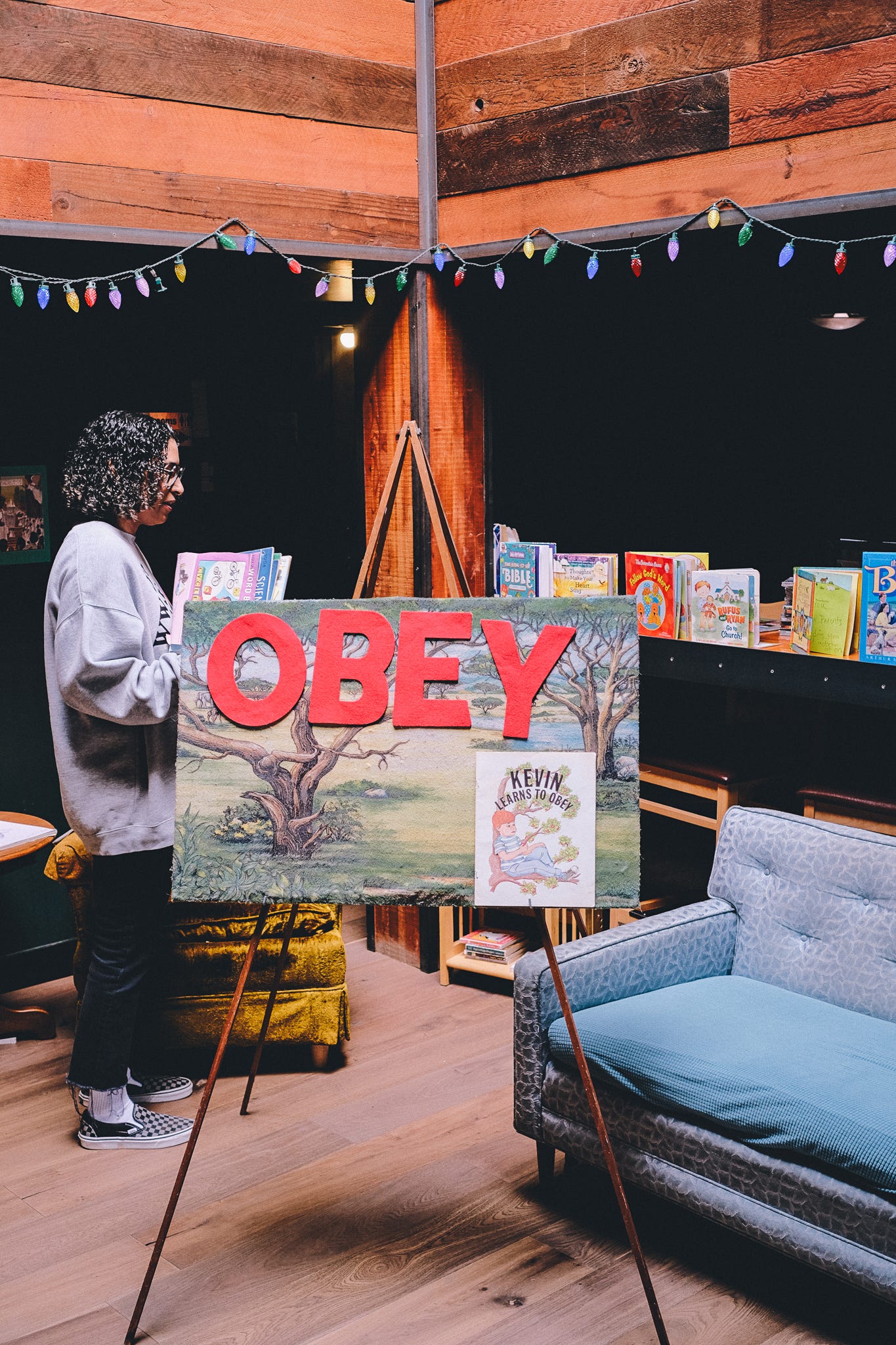 a person with black curly hair is standing in the kid's church corner with a large felt board for large red letters spelling out OBEY on the board with various children's bibles on the shelf in the background