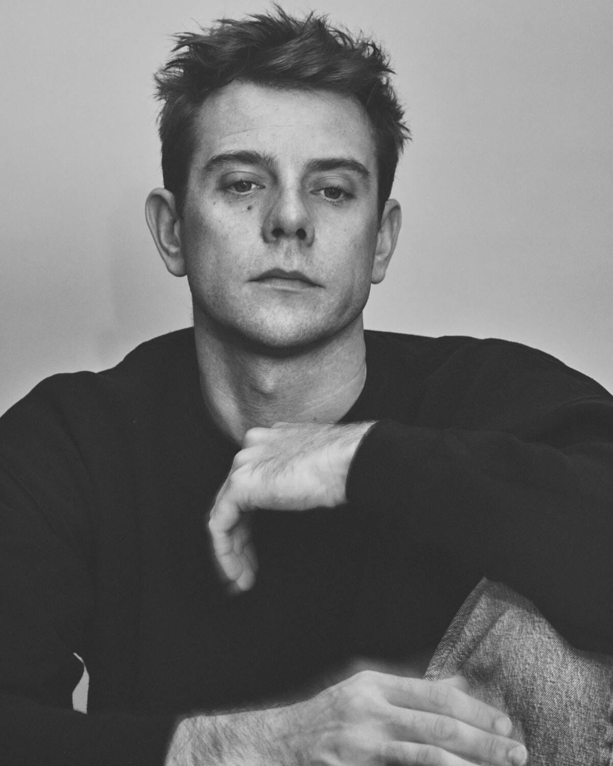 A black and white portrait depicts a young man from the chest up wearing a black sweater and staring off beyond camera.