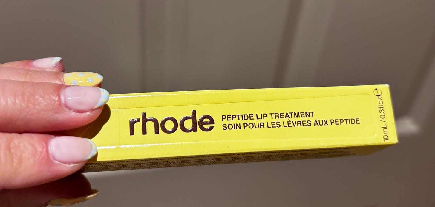 A close-up of the Rhode Peptide Lip Treatment in Caramelised Banana held by a beauty editor, showing the sleek yellow branding.
