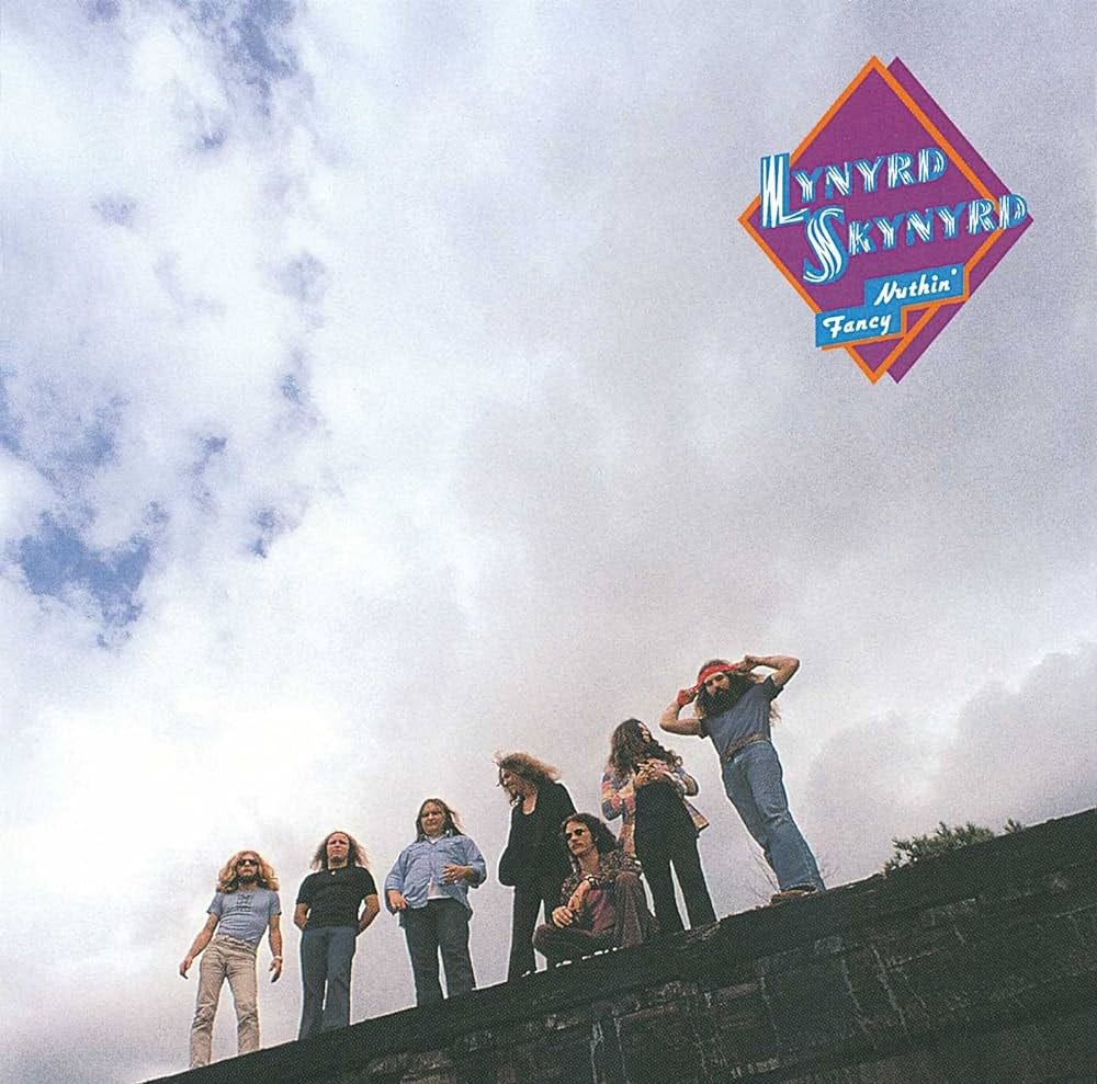 The cover of Lynyrd Skynyrd’s 1975 album. The upper two thirds of the photograph are white clouds and a bit of blue sky in the upper left corner. At the bottom, the severn band members are on top of a wall. Six of them are standing, one of them is squatting. 1970s looks: tight bottom bell pants, tight shirt, long hair, the guy on the right is tying a headband around his head. On the upper right corner, the name of the band and the title of the album, Nuthin’ Fancy, are written in blue and white lettering on a pink and orange square. The cover of Lynyrd Skynyrd’s 1975 album. The upper two thirds of the photograph are white clouds and a bit of blue sky in the upper left corner. At the bottom, the severn band members are on top of a wall. Six of them are standing, one of them is squatting. 1970s looks: tight bottom bell pants, tight shirt, long hair, the guy on the right is tying a headband around his head. On the upper right corner, the name of the band and the title of the album, Nuthin’ Fancy, are written in blue and white lettering on a pink and orange square.