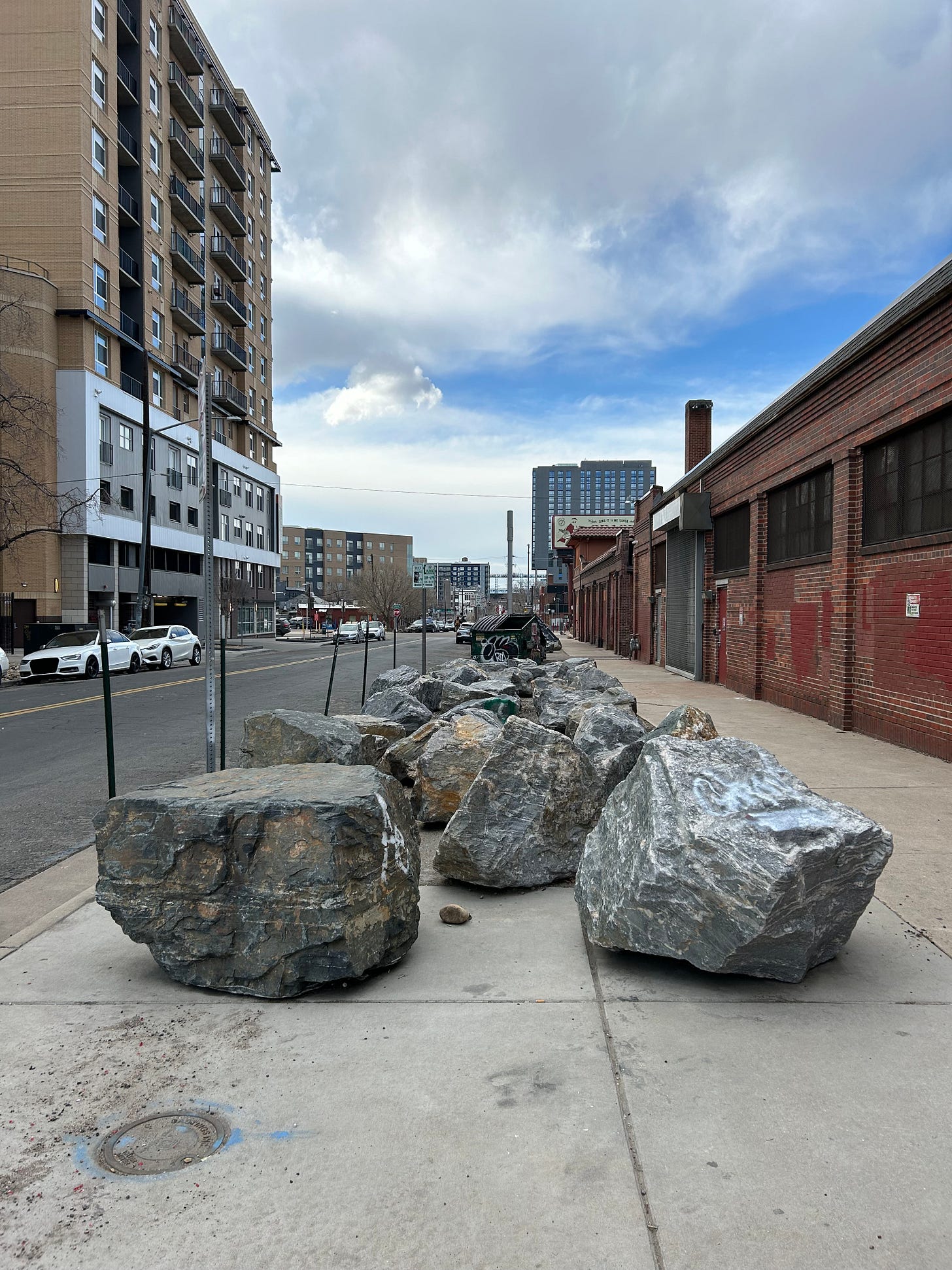 A picture of a streetscape. Half of the sidewalk area closest to the curb is filled with large boulders clustered close together.