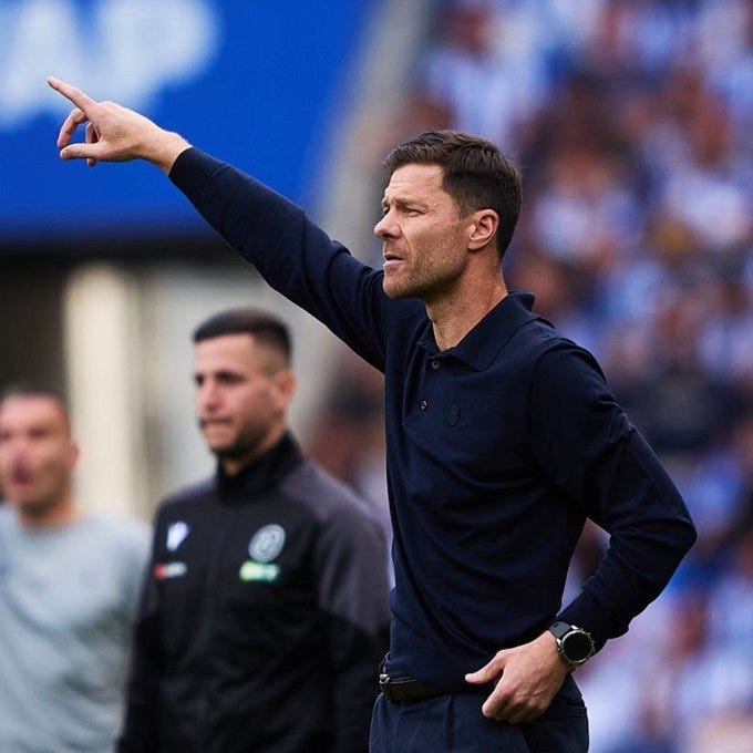 Xabi Alonso stands on a soccer field, pointing with his right arm extended. He wears a dark long-sleeve polo shirt and dark pants, with a watch on his left wrist. Behind him, a man in a black jacket with a logo and another in a gray jacket are visible, with a crowd of spectators in blue and white in the background.